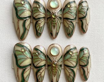 May include: A set of ten press-on nails with a butterfly design. Each nail features a cream-colored base with green and gold accents, creating a stained-glass effect. The butterfly wings are detailed with intricate lines.