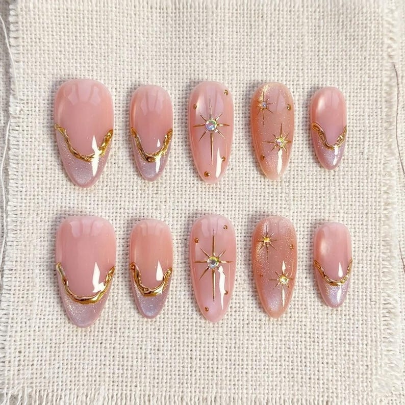 May include: Set of ten artificial nails in various sizes. The nails are oval-shaped with a light pink base and gold accents. Some nails feature a gold starburst design with a central gem, while others have a gold border.