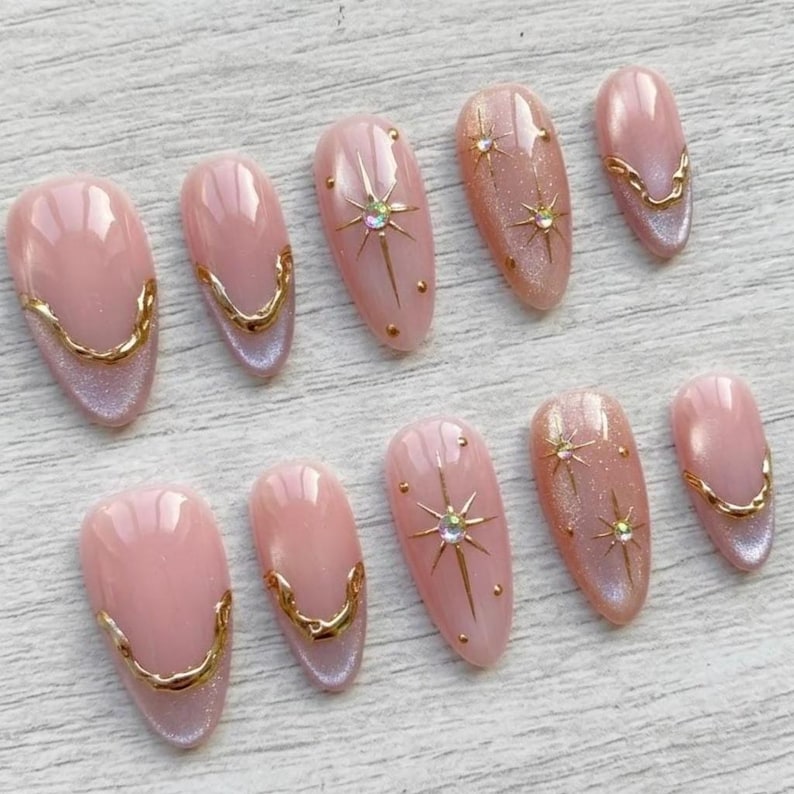 May include: A set of ten artificial nails in various sizes. The nails are oval-shaped with a light pink base and gold accents. Some nails feature gold starburst designs with rhinestones, while others have a gold trim along the base.