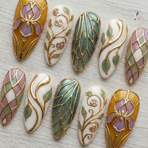 May include: A collection of ten almond-shaped, painted fingernails. Each nail features a unique design with gold metallic outlines and accents. Designs include floral patterns, geometric shapes, and abstract leaf motifs, with colors like white, green, purple, and yellow.