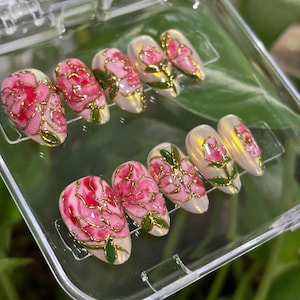 May include: Set of ten artificial nails with a floral design. Each nail features a pink rose with gold outlines and green leaves. The nails are oval-shaped and are displayed in a clear plastic case.