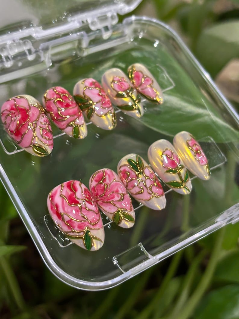 May include: Set of ten artificial nails with a floral design. Each nail features a pink rose with gold outlines and green leaves. The nails are oval-shaped and are displayed in a clear plastic case.