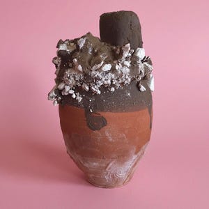 May include: A terracotta-colored ceramic vessel with a tapered base and a wide rim. The upper portion is coated in a thick, dark brown glaze that drips down the sides, topped with a textured cluster of white and brown crumbles and a dark brown cylinder.
