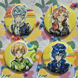 May include: Four round yellow buttons with anime-style character illustrations. Each button features a different character in various outfits, with colorful striped and swirl backgrounds. Small beads are scattered around the buttons.