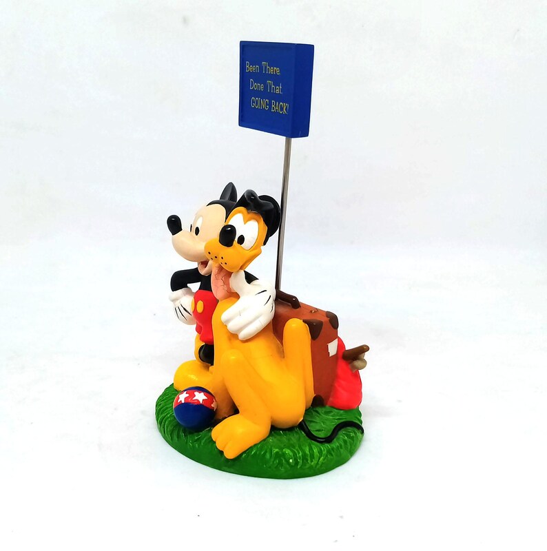 May include: A figurine featuring Mickey Mouse and Pluto embracing, with a blue sign that reads "Been There Done That GOING BACK!". The characters are on a green base with a red, white, and blue ball.
