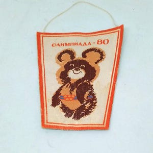 May include: Vintage pennant featuring the Olympic bear Misha, the mascot of the 1980 Moscow Olympics. The pennant has a cream background with a brown bear illustration and the text "ОЛИМПИАДА-80". It has an orange border and a string for hanging.