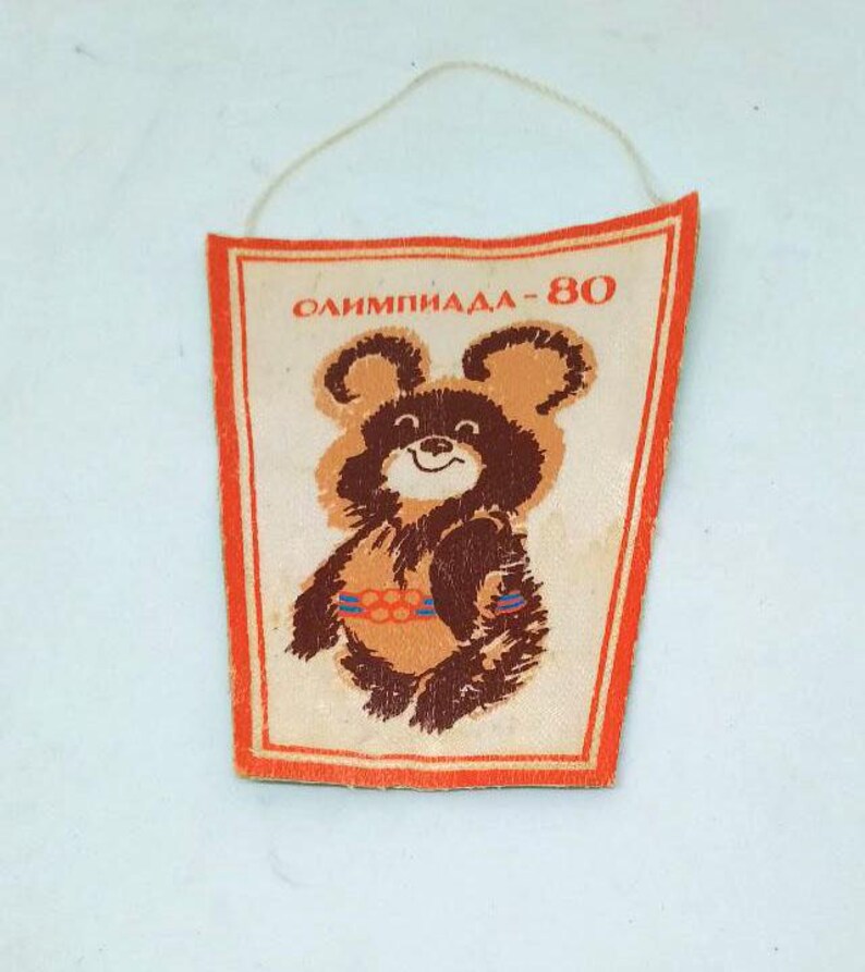 May include: Vintage pennant featuring the Olympic bear Misha, the mascot of the 1980 Moscow Olympics. The pennant has a cream background with a brown bear illustration and the text "ОЛИМПИАДА-80". It has an orange border and a string for hanging.