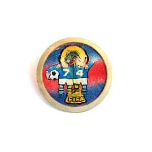 May include: A round, vintage FIFA badge featuring a soccer player in a blue uniform with the number 74. The badge has a white border and a red and blue background. The FIFA logo is at the bottom.