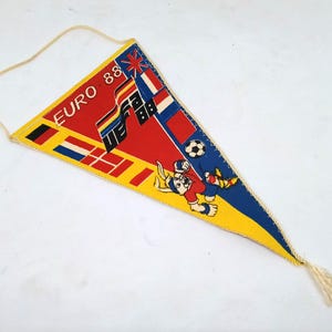 May include: A vintage pennant in a triangular shape, featuring the text "EURO 88" and various flags. The pennant has a yellow, red, and blue color scheme, with a cartoon mascot and a soccer ball. A cream-colored cord is attached.