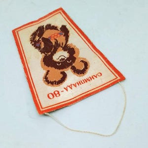 May include: Rectangular vintage patch featuring a brown bear with orange accents, a red border, and the text "ОЛИМПИАДА-80". The bear is depicted in a playful pose. A white string is attached to the patch.