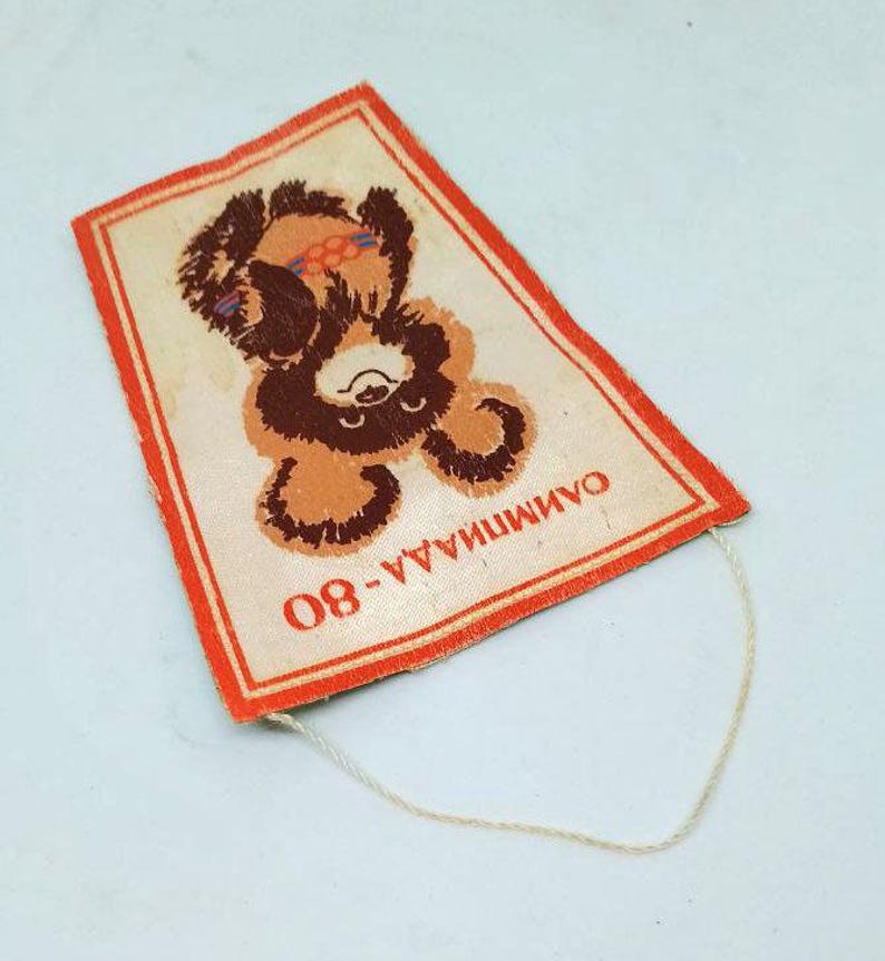 May include: Rectangular vintage patch featuring a brown bear with orange accents, a red border, and the text "ОЛИМПИАДА-80". The bear is depicted in a playful pose. A white string is attached to the patch.