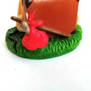 May include: A Disney figurine featuring a brown suitcase, a red sack, and a bone on a green grassy base. The base has the text "&copy; DISNEY CHINA". The figurine is set against a white background.