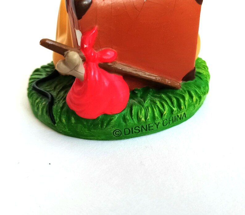 May include: A Disney figurine featuring a brown suitcase, a red sack, and a bone on a green grassy base. The base has the text "&copy; DISNEY CHINA". The figurine is set against a white background.