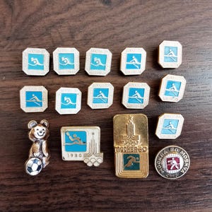May include: A collection of vintage Olympic pins featuring rowing, running, and other sports. The pins are in various shapes and sizes, with blue, white, and gold accents. One pin depicts a bear holding a soccer ball.