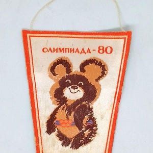 May include: Vintage pennant featuring the Olympic mascot Misha the bear from the 1980 Moscow Olympics. The pennant has a white background with a red border and the text "ОЛИМПИАДА-80". The bear is brown and tan with a smiling face.