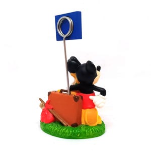 May include: A Mickey Mouse figurine with a blue sign holder. Mickey is dressed in his classic red shorts, yellow shoes, and white gloves, carrying a brown suitcase and a red sack. The base is green with the text "&copy; DISNEY CHINA".
