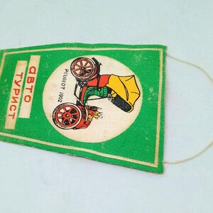 May include: A vintage green pennant with a white circle featuring a red, yellow, and black car illustration. The pennant has the text "ABTO TУРИСТ" in orange and "PEUGEOT 1902" in black. A white string is attached.