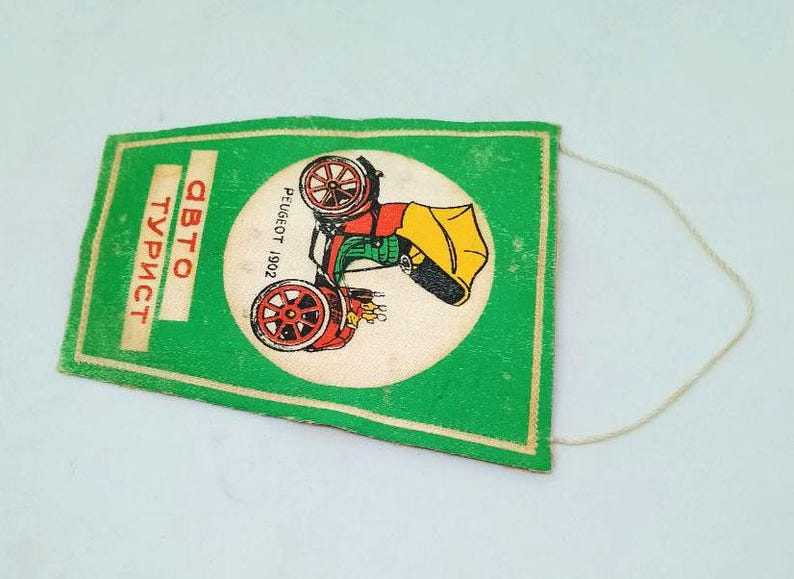 May include: A vintage green pennant with a white circle featuring a red, yellow, and black car illustration. The pennant has the text "ABTO TУРИСТ" in orange and "PEUGEOT 1902" in black. A white string is attached.
