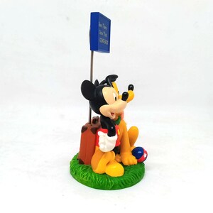 May include: A figurine featuring Mickey Mouse and Pluto sitting on a patch of green grass. Mickey is wearing red shorts and a red shirt, and Pluto is yellow. A blue sign reads "Been There Done That COME BACK". A brown suitcase is next to Mickey.