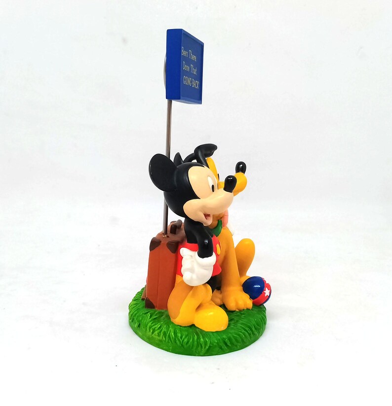 May include: A figurine featuring Mickey Mouse and Pluto sitting on a patch of green grass. Mickey is wearing red shorts and a red shirt, and Pluto is yellow. A blue sign reads "Been There Done That COME BACK". A brown suitcase is next to Mickey.