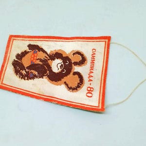 May include: Rectangular vintage tag featuring a brown bear illustration with Olympic rings. The tag has a red border and the text "ОЛИМПИАДА-80" in Russian. A white string is attached to the top right corner.