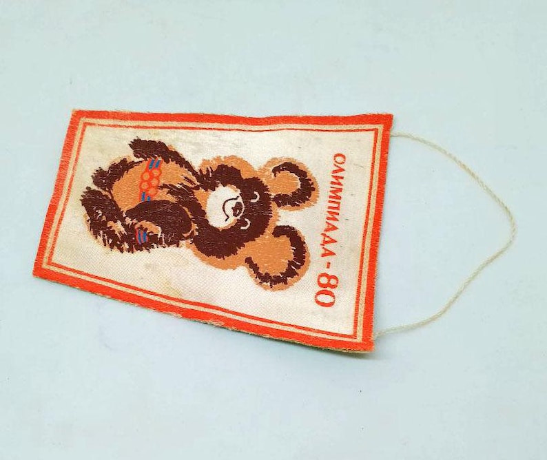 May include: Rectangular vintage tag featuring a brown bear illustration with Olympic rings. The tag has a red border and the text "ОЛИМПИАДА-80" in Russian. A white string is attached to the top right corner.