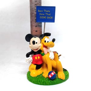 May include: A figurine featuring Mickey Mouse and Pluto, with a blue sign that reads "Been There, Done That, GOING BACK!". Mickey is in his classic outfit, and Pluto is yellow with a pink tongue. A red, white, and blue ball sits on the green base.