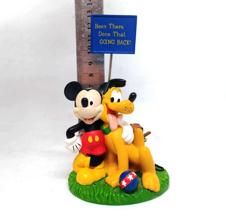May include: A figurine featuring Mickey Mouse and Pluto, with a blue sign that reads "Been There, Done That, GOING BACK!". Mickey is in his classic outfit, and Pluto is yellow with a pink tongue. A red, white, and blue ball sits on the green base.