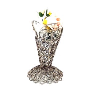 May include: A silver-toned metal vase featuring an intricate, openwork filigree design with a flared, scalloped rim and a wide, circular base. The vase contains a small arrangement of artificial flowers with white and pale pink petals.