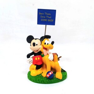 May include: A figurine featuring Mickey Mouse and Pluto embracing on a green base. Mickey wears red shorts and Pluto is yellow. A blue sign above reads "Been There. Done That. GOING BACK!" A red, white, and blue ball is on the base.