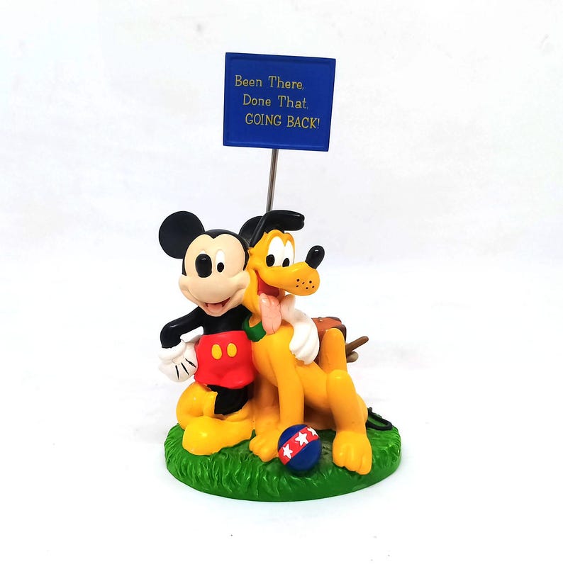 May include: A figurine featuring Mickey Mouse and Pluto embracing on a green base. Mickey wears red shorts and Pluto is yellow. A blue sign above reads "Been There. Done That. GOING BACK!" A red, white, and blue ball is on the base.