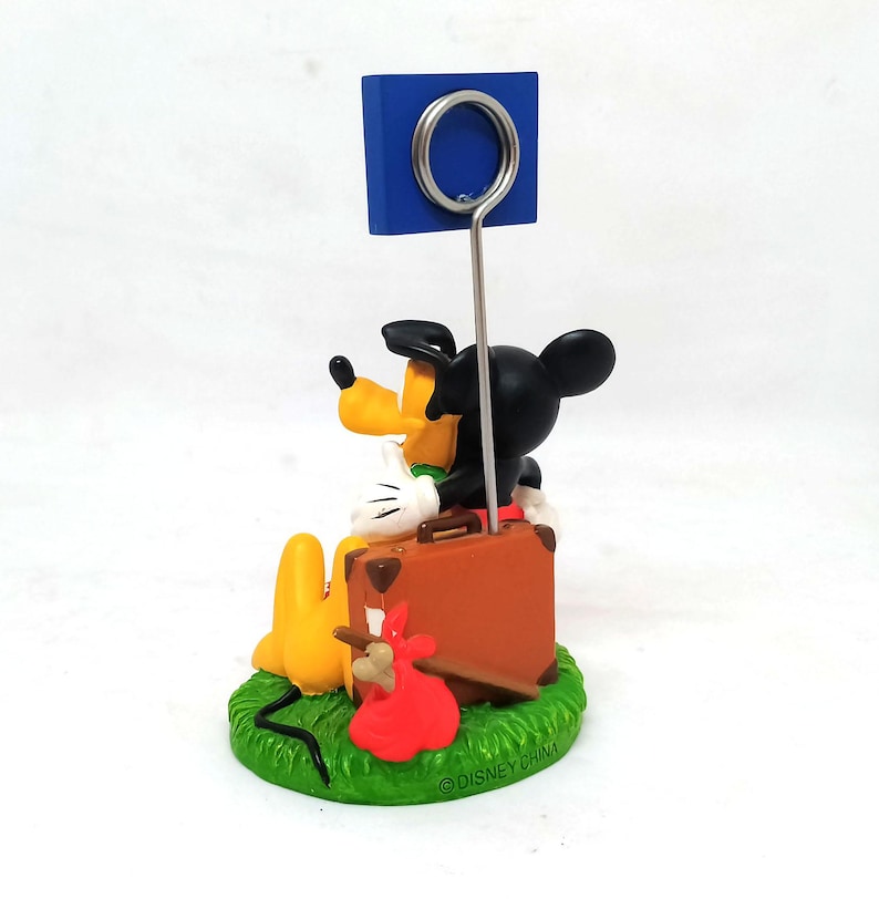 May include: A whimsical Disney-themed photo holder featuring Pluto and Mickey Mouse. Pluto is lying on a green base, while Mickey sits on a brown suitcase. A blue square holds a metal ring for displaying photos.