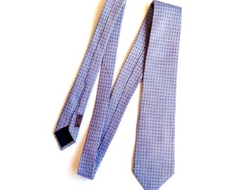 May include: A light blue necktie featuring a repeating geometric pattern of small white squares and dark blue dots. The tie is laid out flat, showing the pointed blade, the folded body, and the dark fabric label on the underside of the narrow end.