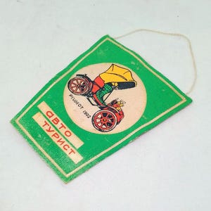 May include: Vintage green tag with a cream-colored circle featuring a colorful illustration of a Peugeot 1902 car. The tag has the words "Авто Турист" in red and white text. A string is attached to the top.