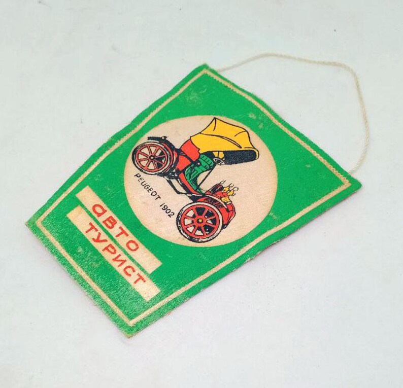 May include: Vintage green tag with a cream-colored circle featuring a colorful illustration of a Peugeot 1902 car. The tag has the words "Авто Турист" in red and white text. A string is attached to the top.