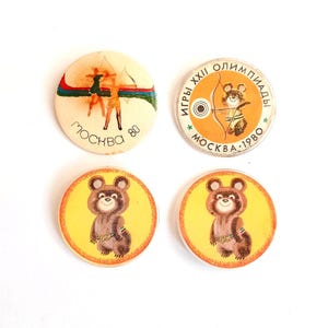 May include: Four vintage Moscow 1980 Olympic pins. Two pins feature the Olympic bear mascot, and two pins feature archers. The pins are round with a cream, yellow, and orange color scheme.