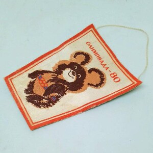 May include: A vintage rectangular pennant featuring the Olympic bear mascot. The bear is brown and tan, holding colorful rings. The pennant has a red border and the text "Олимпиада-80" in red. A string is attached for hanging.