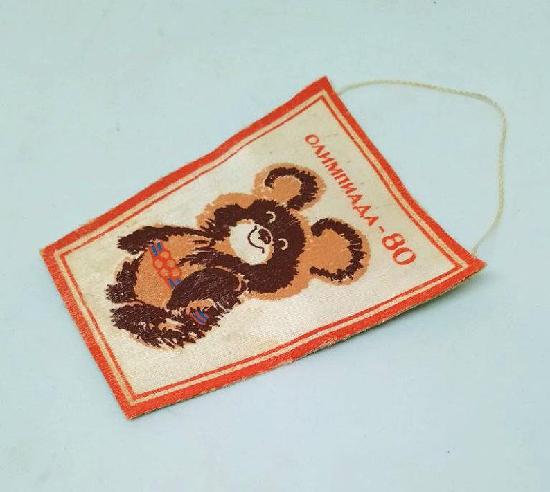 May include: A vintage rectangular pennant featuring the Olympic bear mascot. The bear is brown and tan, holding colorful rings. The pennant has a red border and the text "Олимпиада-80" in red. A string is attached for hanging.