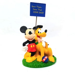 May include: A figurine featuring Mickey Mouse and Pluto, with a blue sign that reads "Been There, Done That, Going Back!" The characters are on a green base with a red, white, and blue ball.