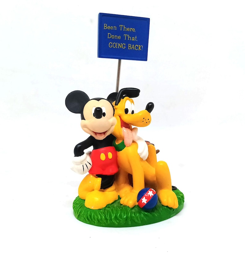 May include: A figurine featuring Mickey Mouse and Pluto, with a blue sign that reads "Been There, Done That, Going Back!" The characters are on a green base with a red, white, and blue ball.