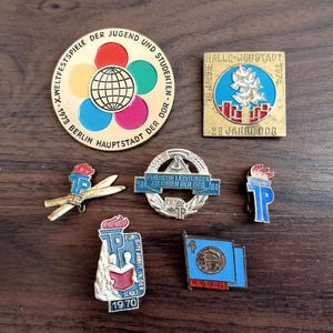 May include: A collection of vintage pins and badges, featuring various designs and text. The pins include a gold-colored pin with a globe and colorful circles, and a rectangular pin with text and a blue and red design. Other pins have torches and flags.