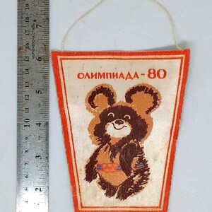 May include: Vintage pennant featuring the Olympic mascot, a brown bear with a smiling face. The pennant has the text "ОЛИМПИАДА-80" in red. The pennant is rectangular with a red border and a string for hanging.