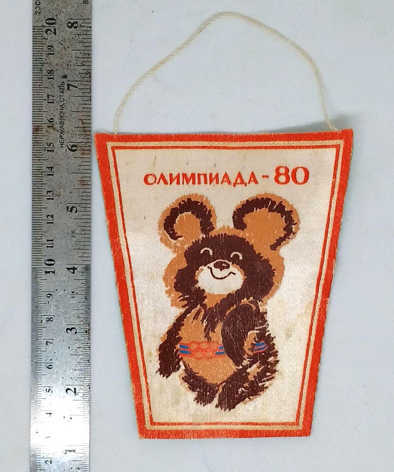 May include: Vintage pennant featuring the Olympic mascot, a brown bear with a smiling face. The pennant has the text "ОЛИМПИАДА-80" in red. The pennant is rectangular with a red border and a string for hanging.