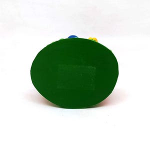May include: A green, oval-shaped decorative item with a flat base. The top edge is textured and has small blue and yellow accents. The item is set against a white background.