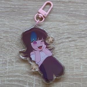 May include: A clear acrylic keyring featuring a smiling anime-style character with brown hair, a blue streak, and a pink eye. The character wears a white top and purple trousers. The keyring has a pink clasp.