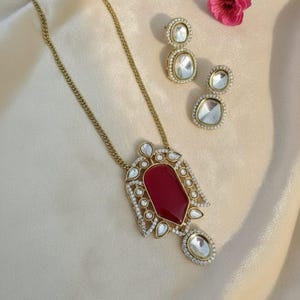 May include: A gold-toned necklace with a large, red, gemstone pendant and matching earrings. The pendant and earrings are adorned with clear, sparkling accents. The jewelry set is displayed on a cream-colored surface.