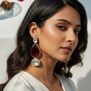 Peut inclure: Boucles d'oreilles pendantes élégantes avec une pierre supérieure transparente de forme carrée, une grande pierre centrale rouge rubis en forme de goutte et une pierre inférieure transparente de forme ronde. Serties dans du métal doré, elles sont entourées de petits cristaux transparents.
