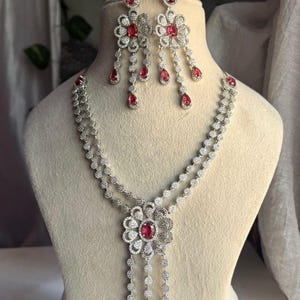 May include: A silver-toned jewelry set featuring a necklace and matching earrings. The necklace has a floral pendant with a large red gemstone and dangling teardrop-shaped gems. The earrings mirror the pendant design, with red gems and dangling accents.