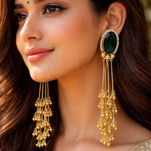 May include: Gold-toned dangle earrings with a large, oval, emerald-green stone surrounded by small pearls. The earrings have multiple strands of gold chains ending in small gold beads, creating a cascading effect. The earrings are a statement piece.