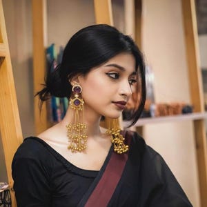 May include: Gold-toned earrings with a dangling design. Each earring features a purple gemstone at the top, with multiple strands of small gold beads cascading downwards. The earrings are worn with a black top.